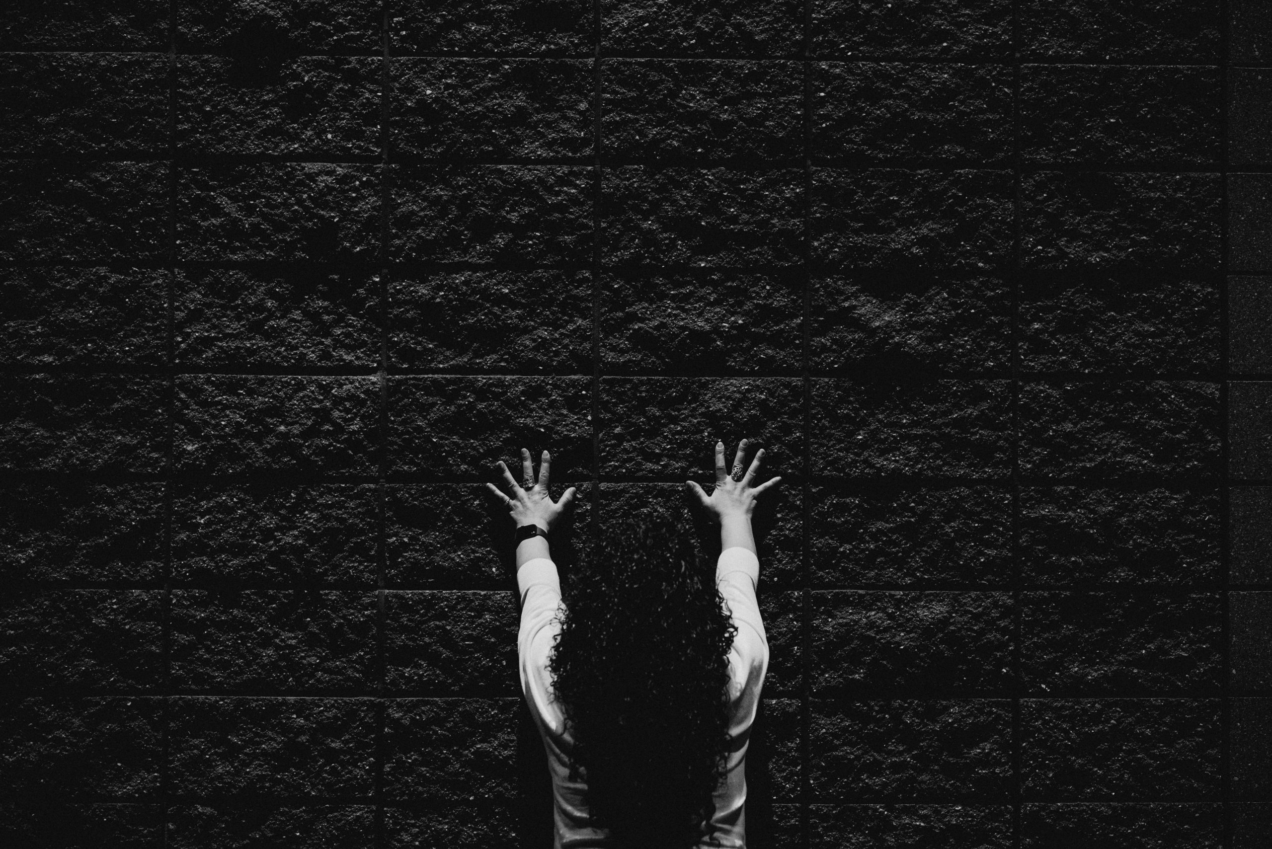 Photo en noir et blanc d’une personne face à un mur de briques sombre, les bras levés, doigts écartés.