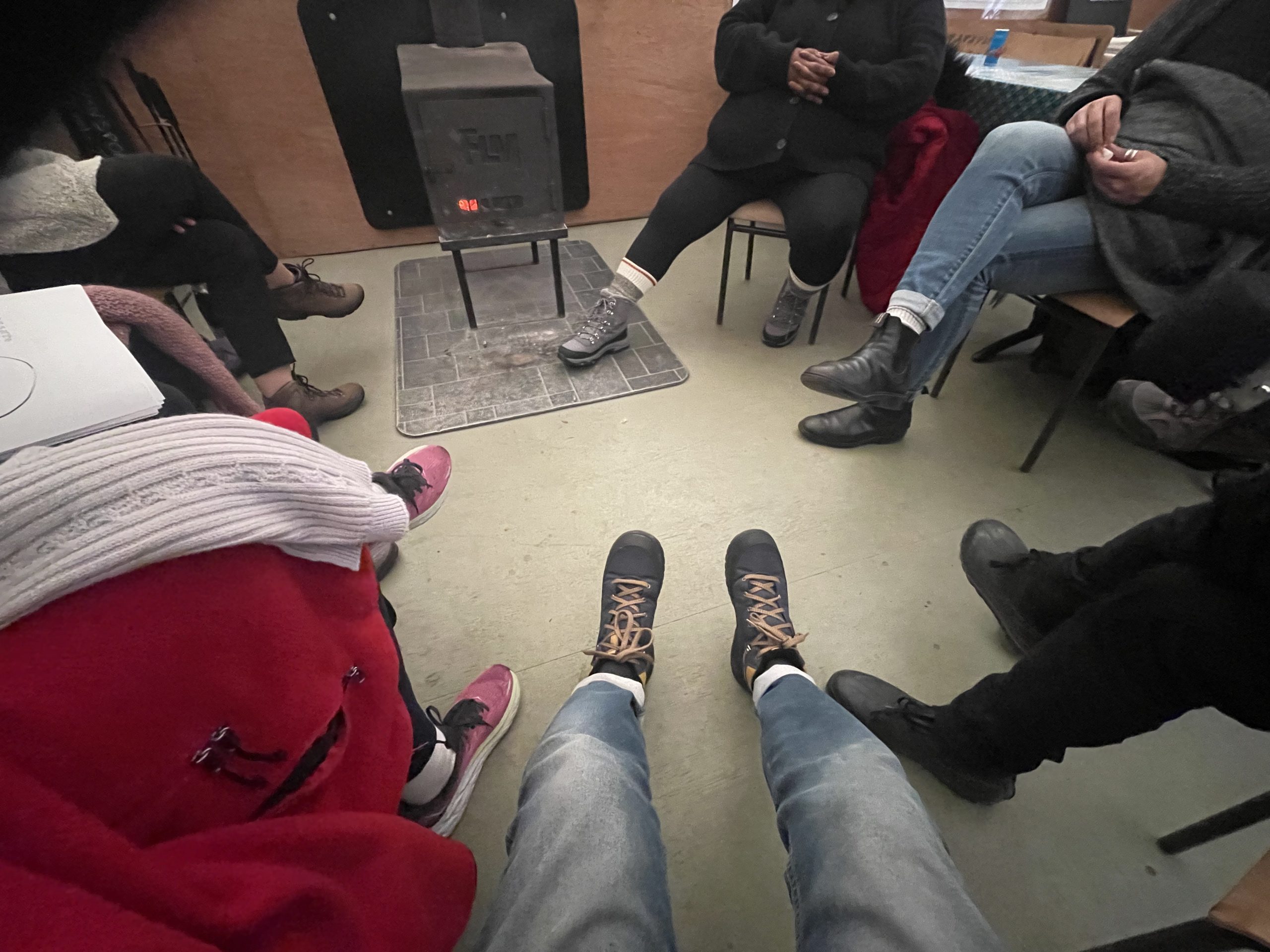 Vue à la première personne de pieds au centre d’un cercle de personnes assises autour d’un poêle à bois.