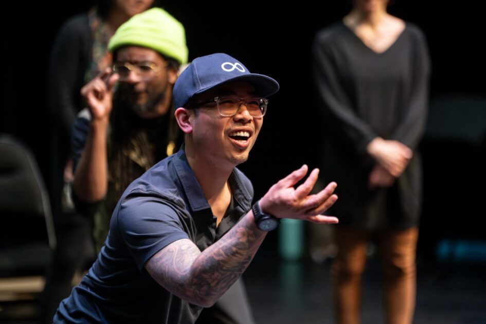 An Asian man wearing a baseball cap is in performance and gesticulates to the crowd.