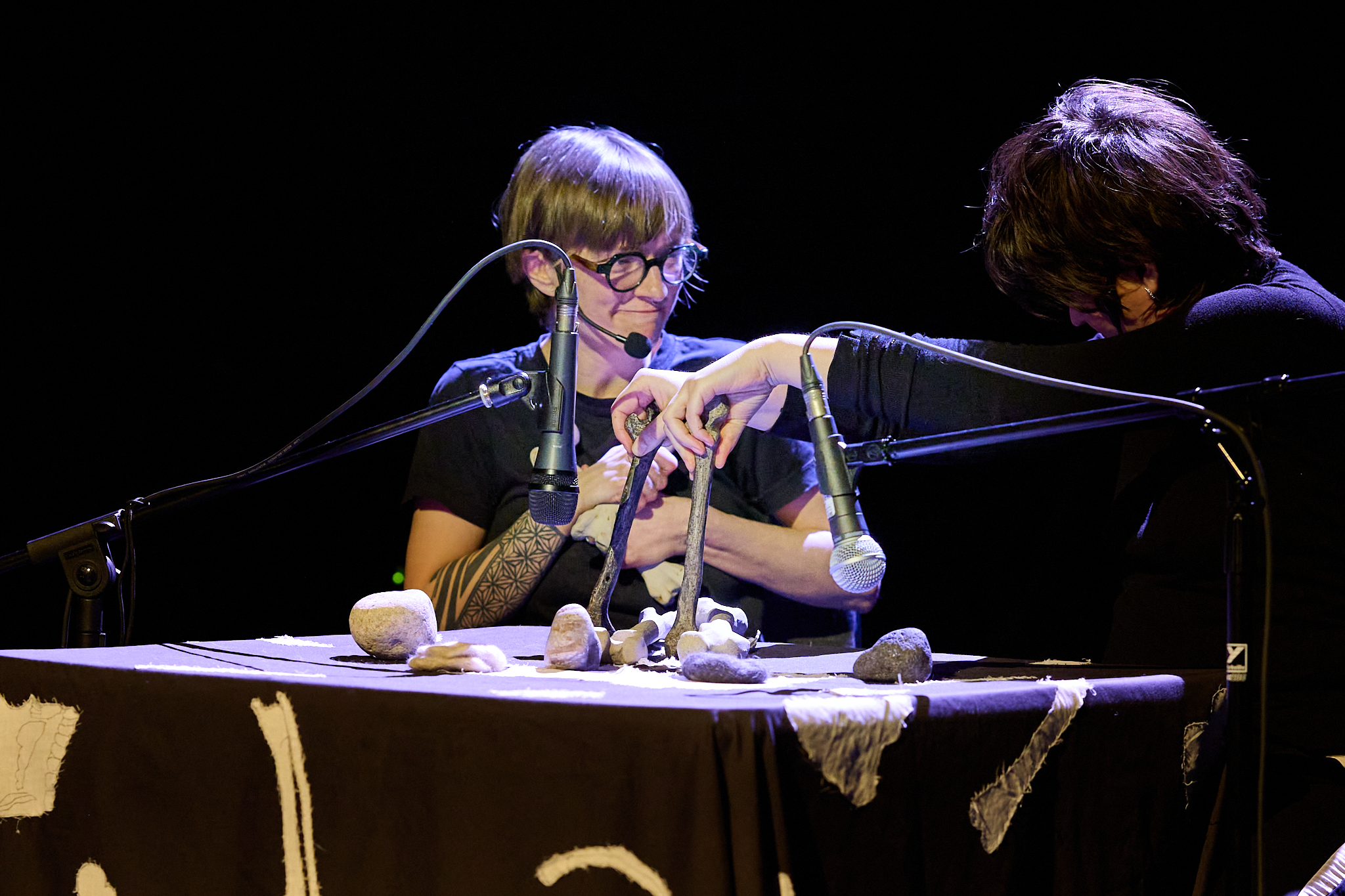 Two performers at a table with microphones; one arranges small objects while the other leans in close.