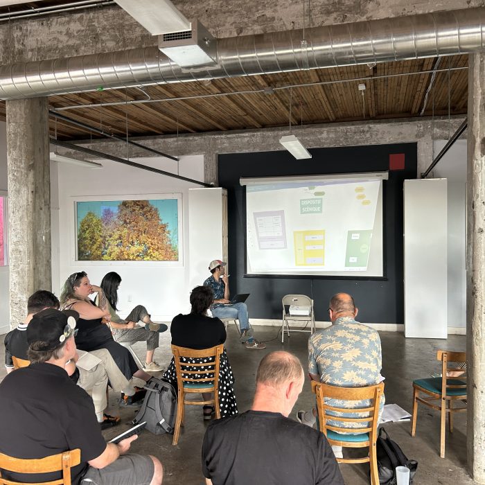 A group of people in a circle are looking at a slide projection in a warehouse space. We see them from behind, observing, looking at their notes.