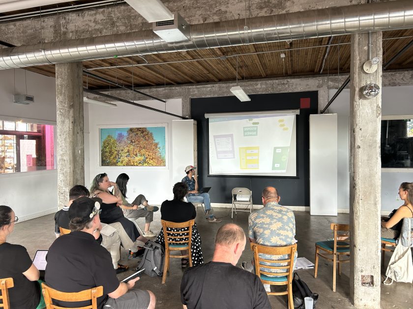 A group of people in a circle are looking at a slide projection in a warehouse space. We see them from behind, observing, looking at their notes.