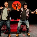 Two men on a stage gesticulate towards an audience as they host a performance cabaret.