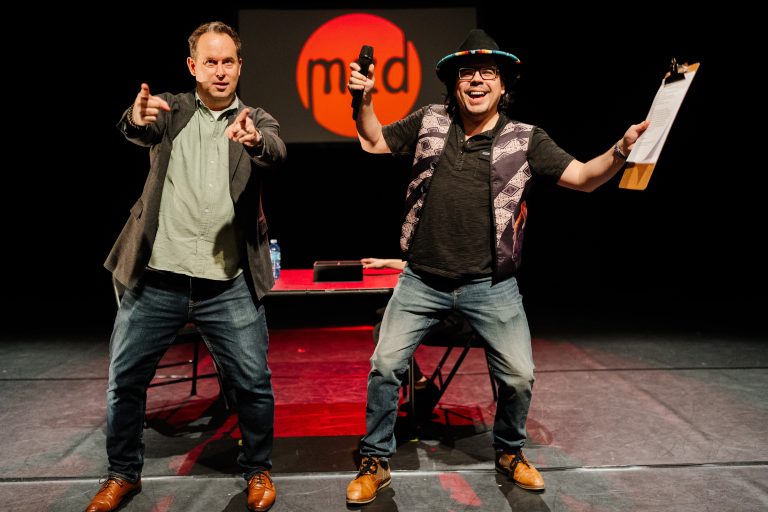 Two men on a stage gesticulate towards an audience as they host a performance cabaret.
