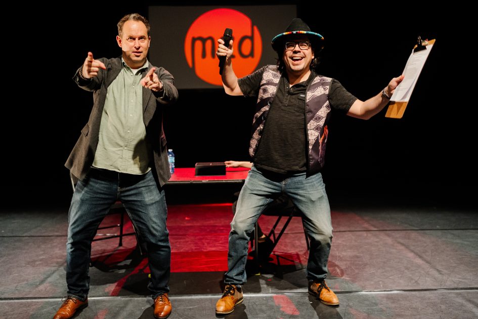 Two men on a stage gesticulate towards an audience as they host a performance cabaret.