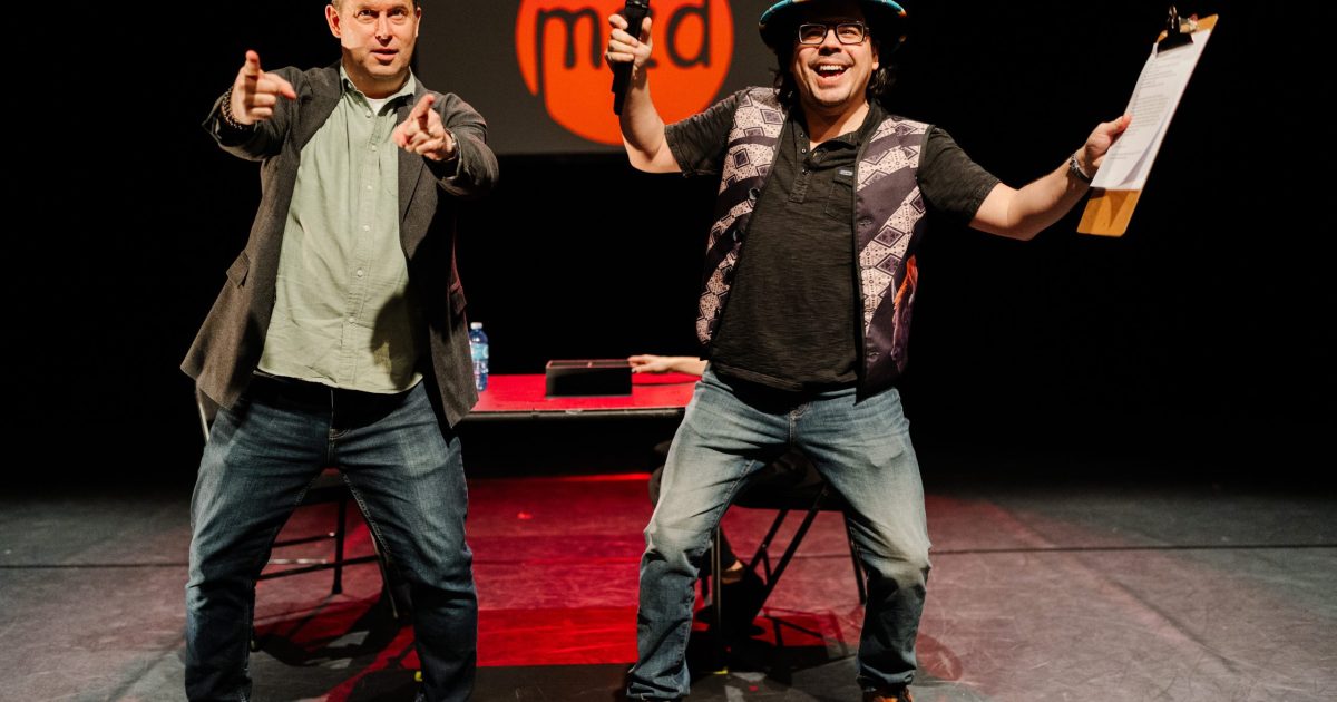 Two men on a stage gesticulate towards an audience as they host a performance cabaret.