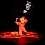 A performer with a bag over their head and dressed in orange kneels on a painted tapestry on the floor. There is a video of a man above them.