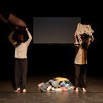 Two performers on either side of a pile of clothing raise shirts over their heads.
