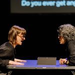 Two animated performers dressed in black face each other with angry expressions at opposite sides of a table. Text projected on the screen behind them reads "Do you ever get angry"