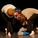 A pair of performers huddle over a pile of clothing attempting to lift it from the floor.
