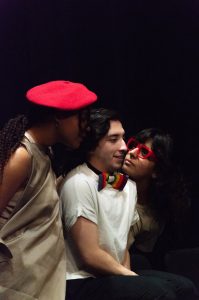 A man with brown hair is surrounded by two women. One of the women wears a red hat and the other red glasses.