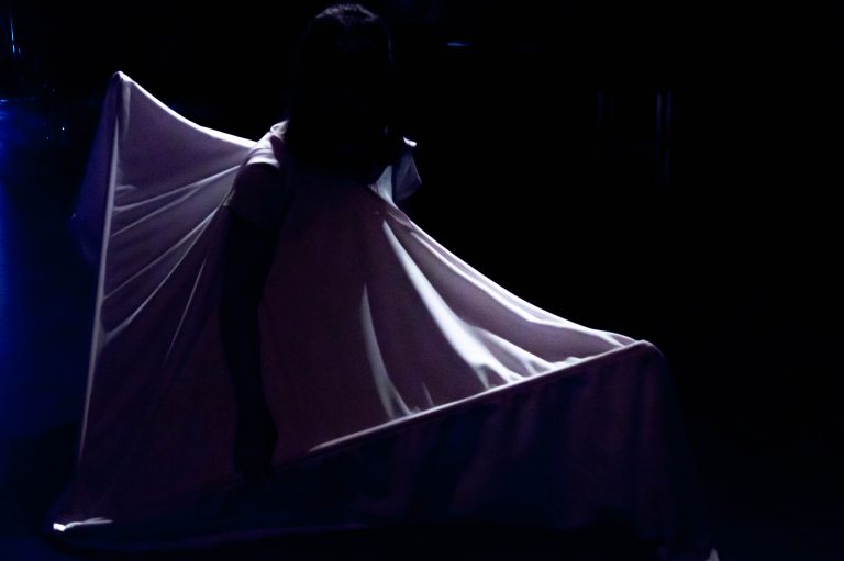 A figure wears a large sail like cloth which is illuminated in a stage light