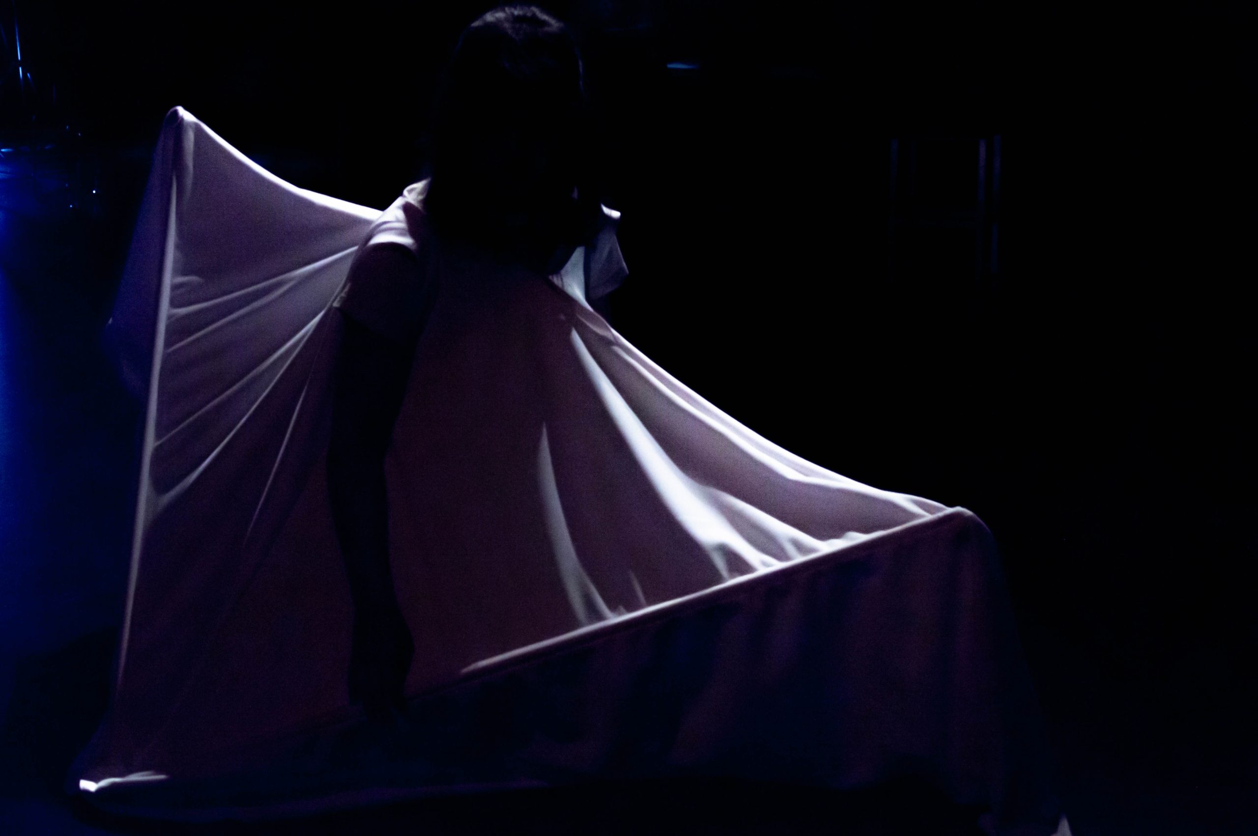 A figure wears a large sail like cloth which is illuminated in a stage light