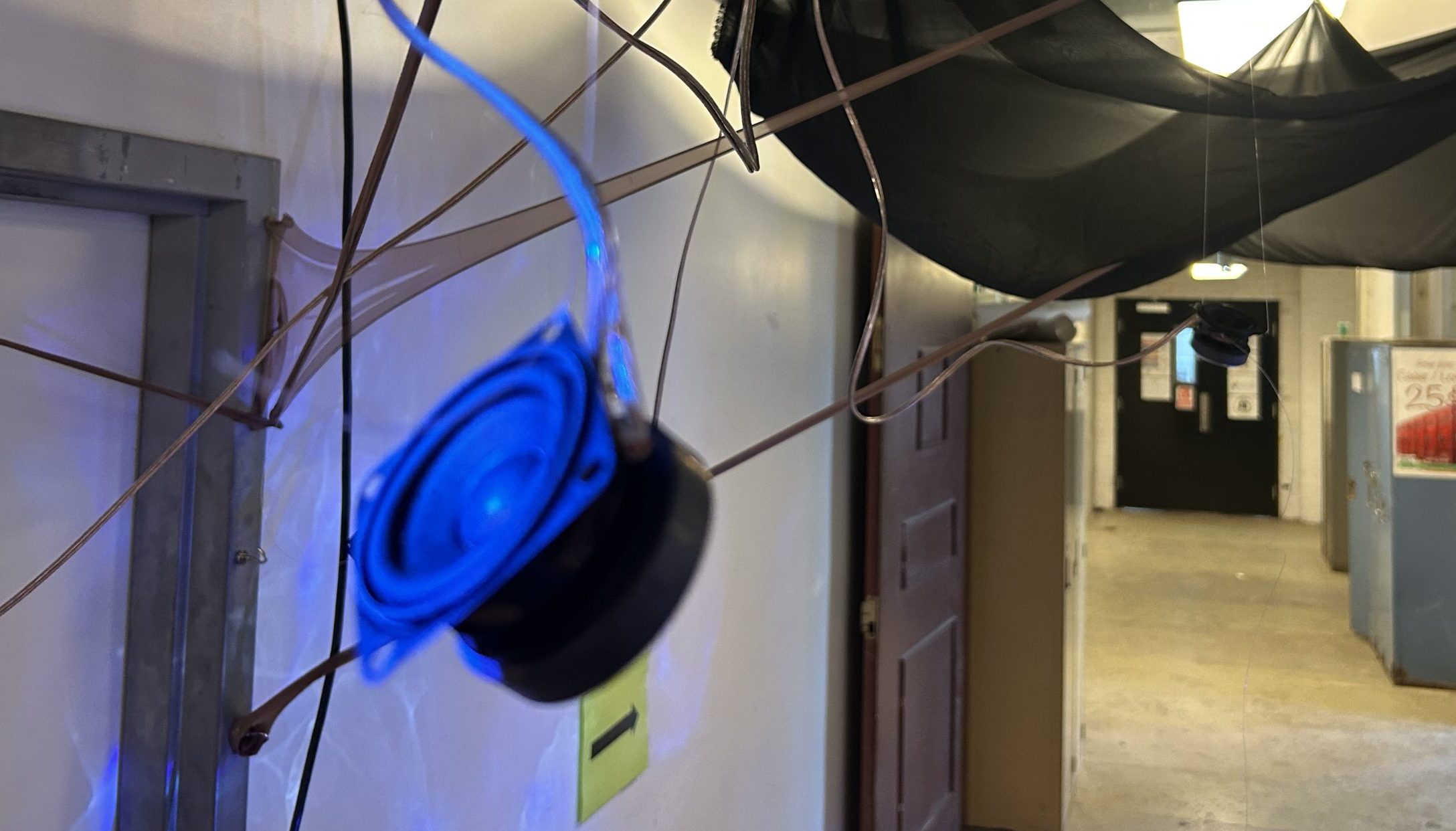 In a building hallway, the image is forgrounded by a speaker, hanging from a wire, and cloth that is stretched over the ceiling.