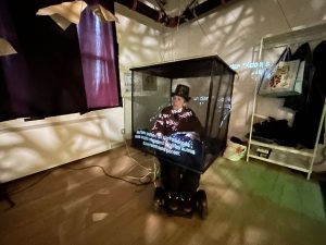 A woman in a wheelchair is covered in a black transluscent box. White text is projected onto the box.