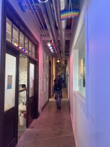 A man walks with his back to the camera down a long hallway filled with entranceways and windows. 