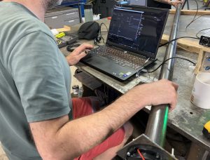 In an industrial setting, a man holds a metal pipe in one hand and types in a computer with the other. 