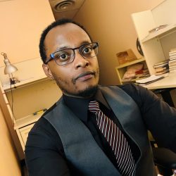 A dark-skinned man with a small beard and glasses looks offscreen at a camera in an office environment. He is dressed in a dark grey vest, black shirt and stripped purple tie.